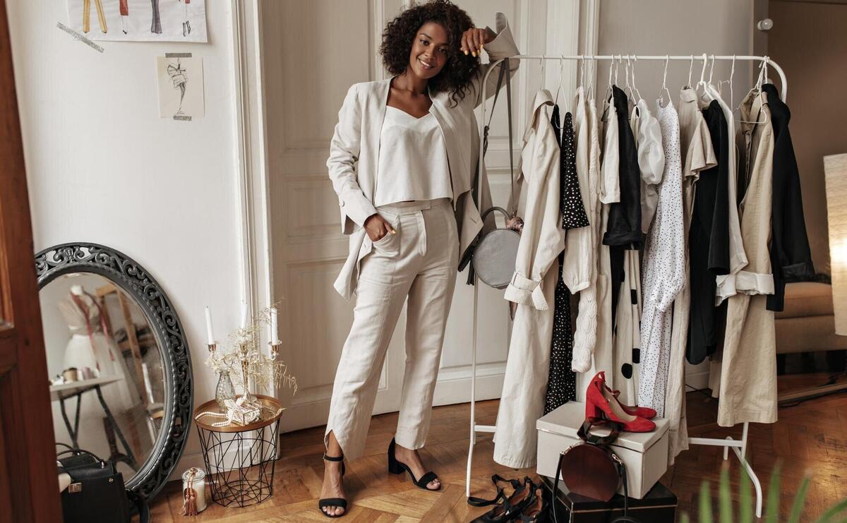 Fulllength portrait of stylish darkskinned curly woman in white pants jacket and top posing in dressing room Young lady leans on hanger