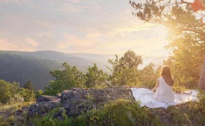Woman in white dress meditating on a rocky cliff during sunset with forested hills in the background